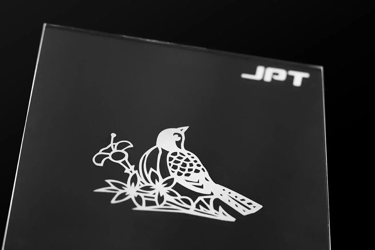 Lasergravur auf einer Glasplatte. Ein Vogel sitzt auf einer Blumenwiese.